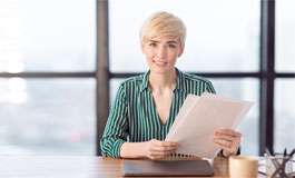 woman sitting at desk, looking at camera with confidence, holding paperwork